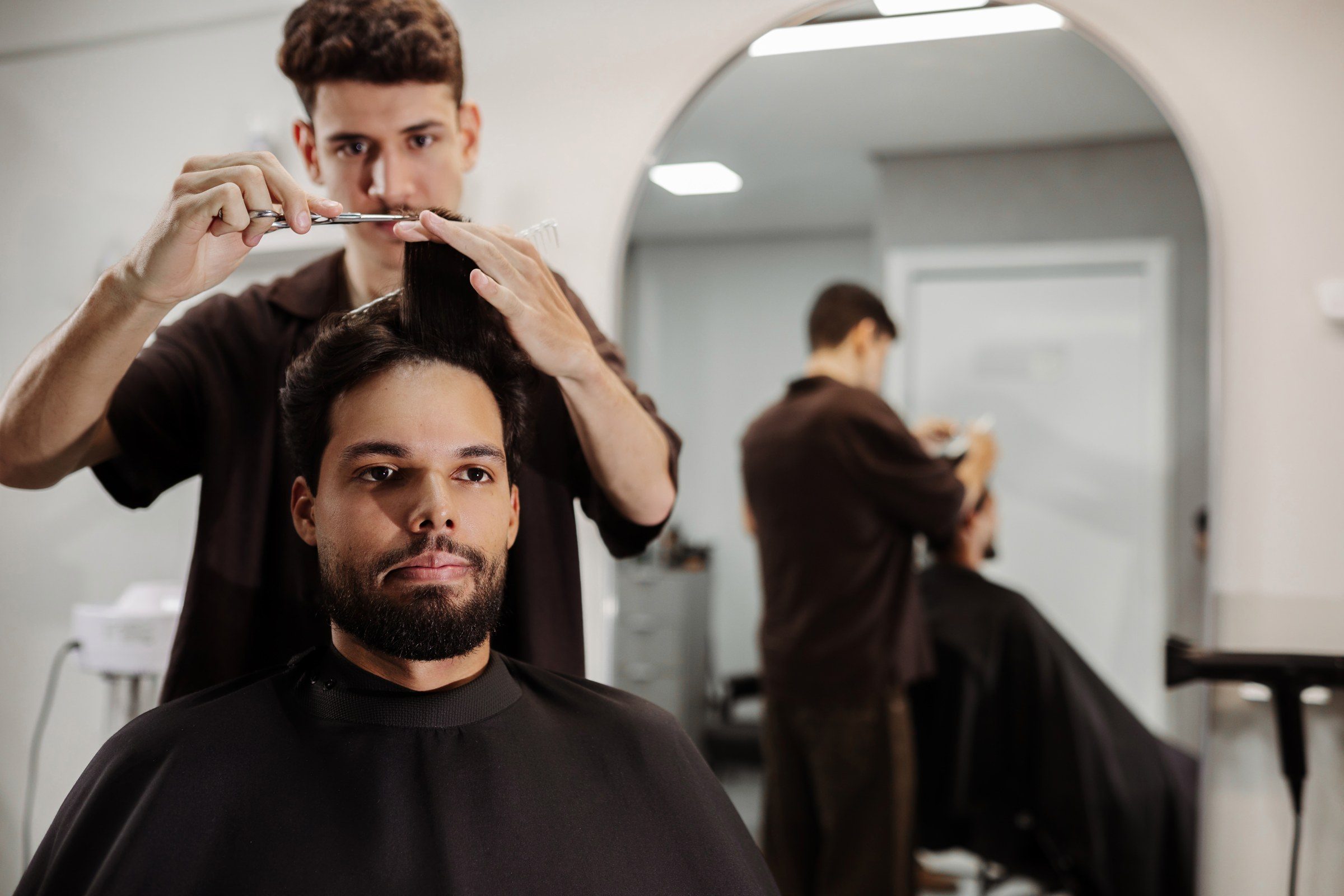 Gabriel cortando o cabelo de um cliente com tesoura, em frente ao espelho em arco da barbearia