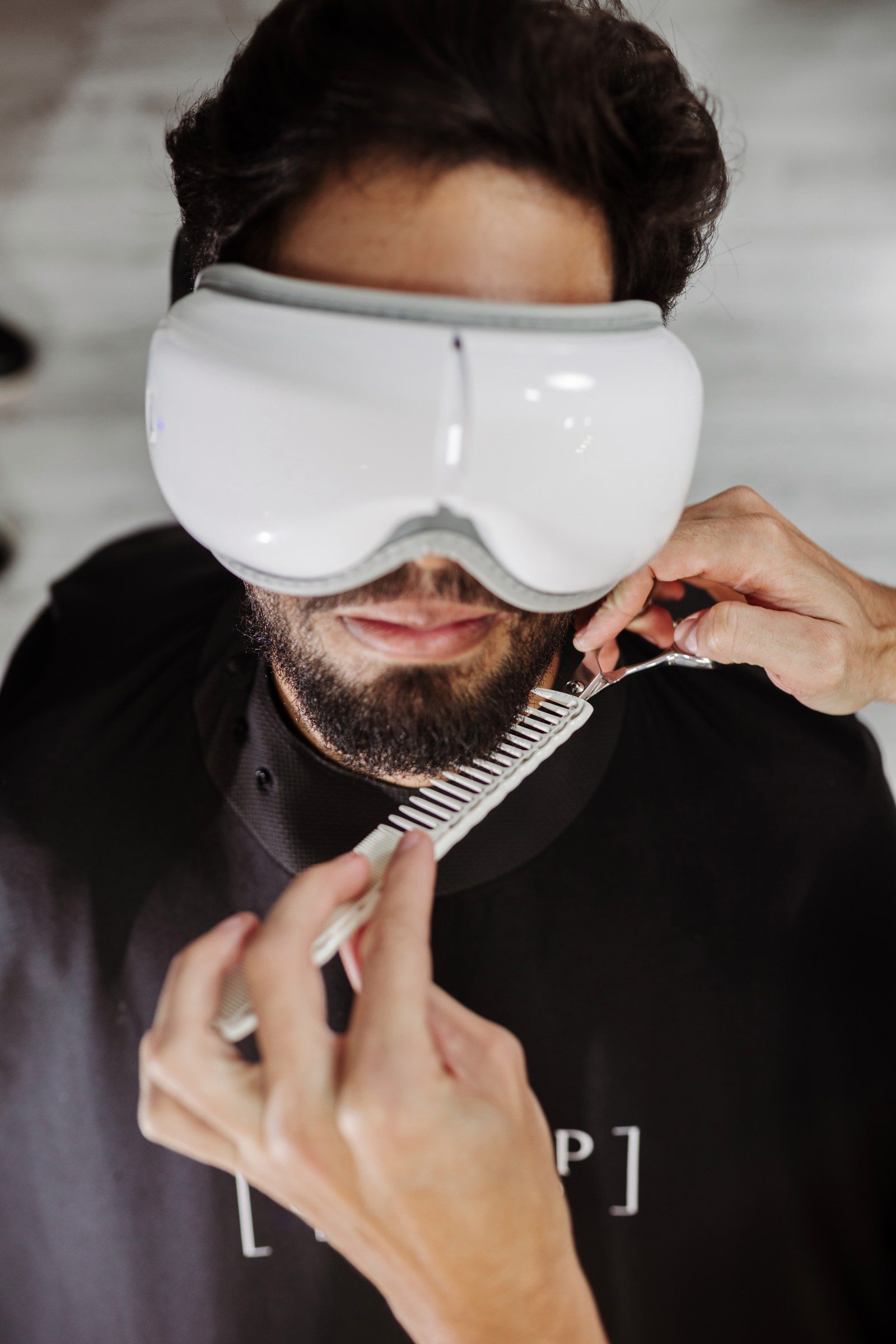 Cliente com massageador ocular enquanto a barba é aparada com tesoura e pente